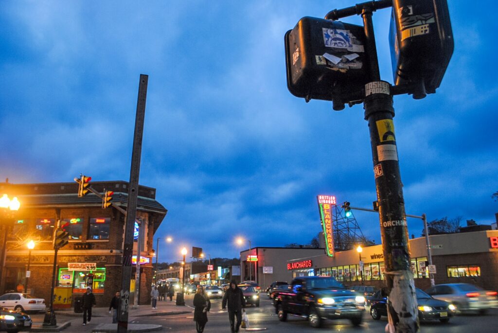 Harvard Avenue in Allston at dusk with lit storefronts and the Blanchards neon sign