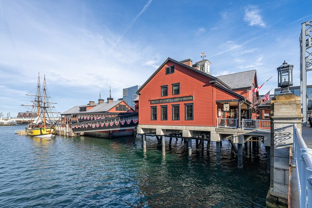 The red Boston Tea Party Ships and Museum building perched over Fort Point Channel at the Congress Street Bridge, with a replica tall ship docked alongside under a blue sky