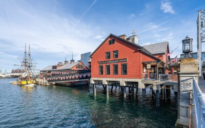 The red Boston Tea Party Ships and Museum building perched over Fort Point Channel at the Congress Street Bridge, with a replica tall ship docked alongside under a blue sky