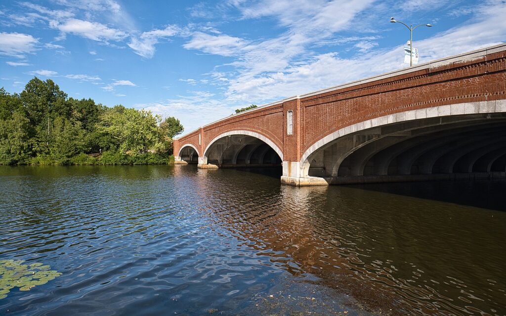 Brick arches of the Eliot Bridge spanning the Charles River beside leafy riverbanks on a sunny summer day