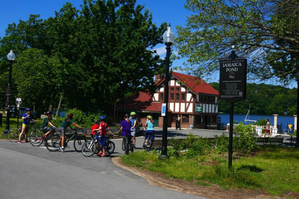 Cyclists by the Emerald Necklace Jamaica Pond sign with the 1892 Tudor boathouse and pond behind