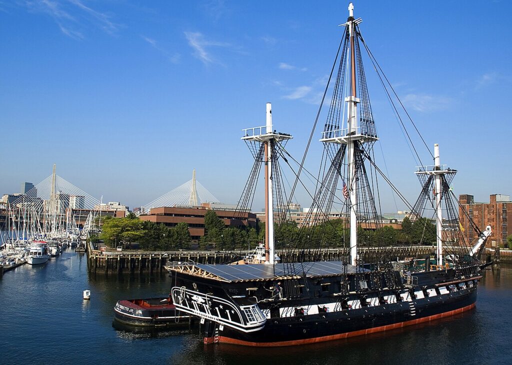 USS Constitution moored at the Charlestown Navy Yard under a clear blue sky, with the Zakim Bridge visible in the background